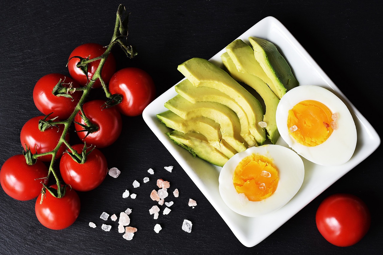 Overhead photo of a white plate with sliced avocado and halved hard-boiled eggs, beside a vine of cherry tomatoes and a few crystals of coarse salt on dark slate.